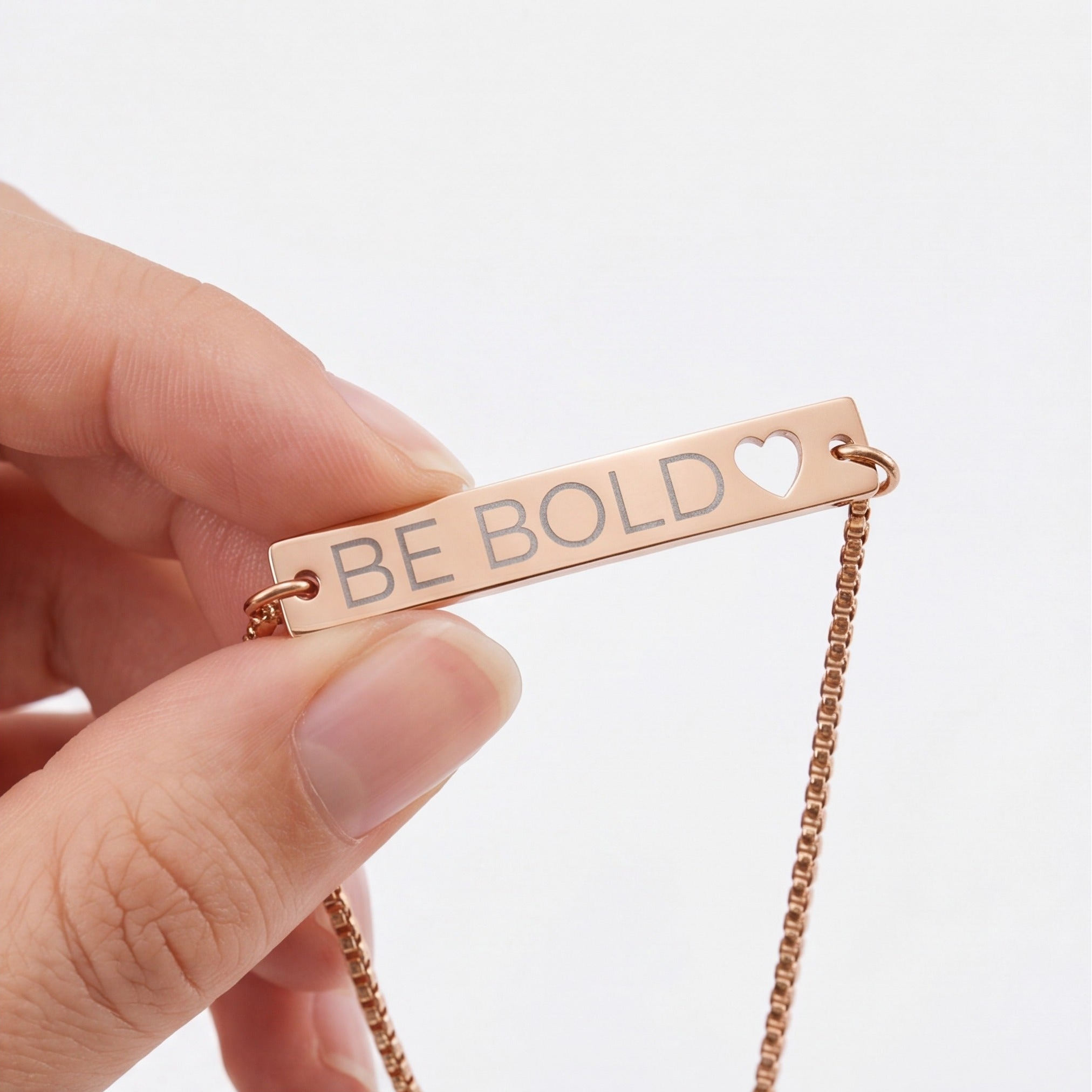 Rose gold necklace with 'BE BOLD' pendant held by a hand on a light background